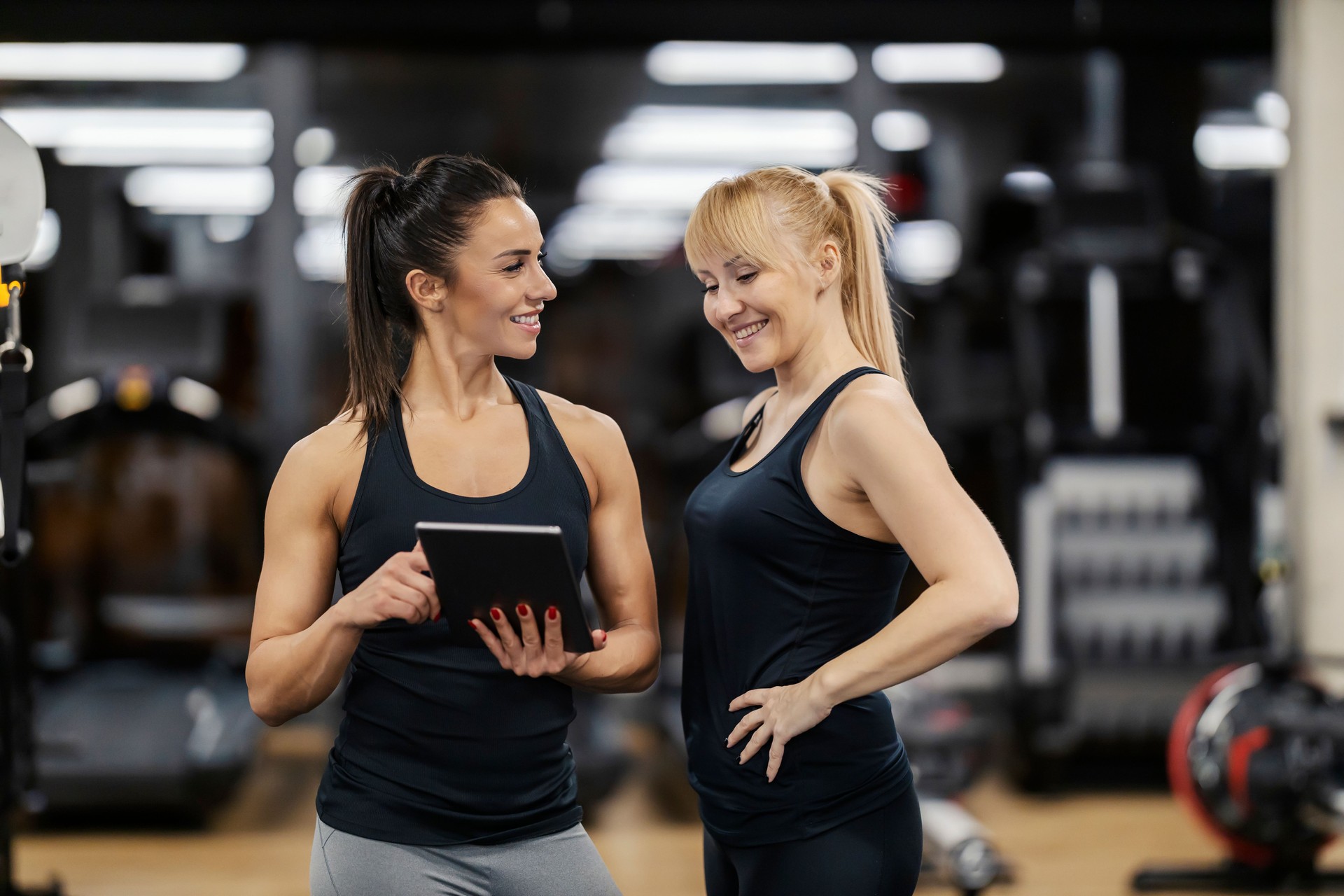 A happy sportswoman is looking at her progress on tablet while female coach is pointing at the tablet.